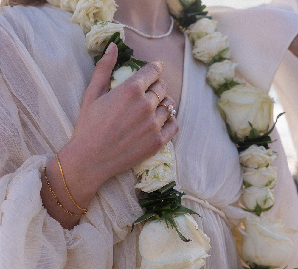 Zarte, goldene Armbänder und Ring von PURELEI, umgeben von Rosenblüten, elegant und feminin.